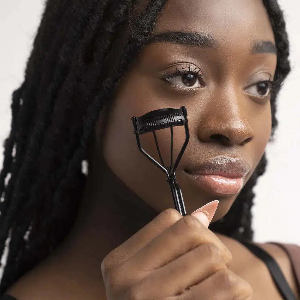 Close-up of a Lashy Cosmetics model with eyes opened, holding a black Lashy eyelash curler against her cheek.